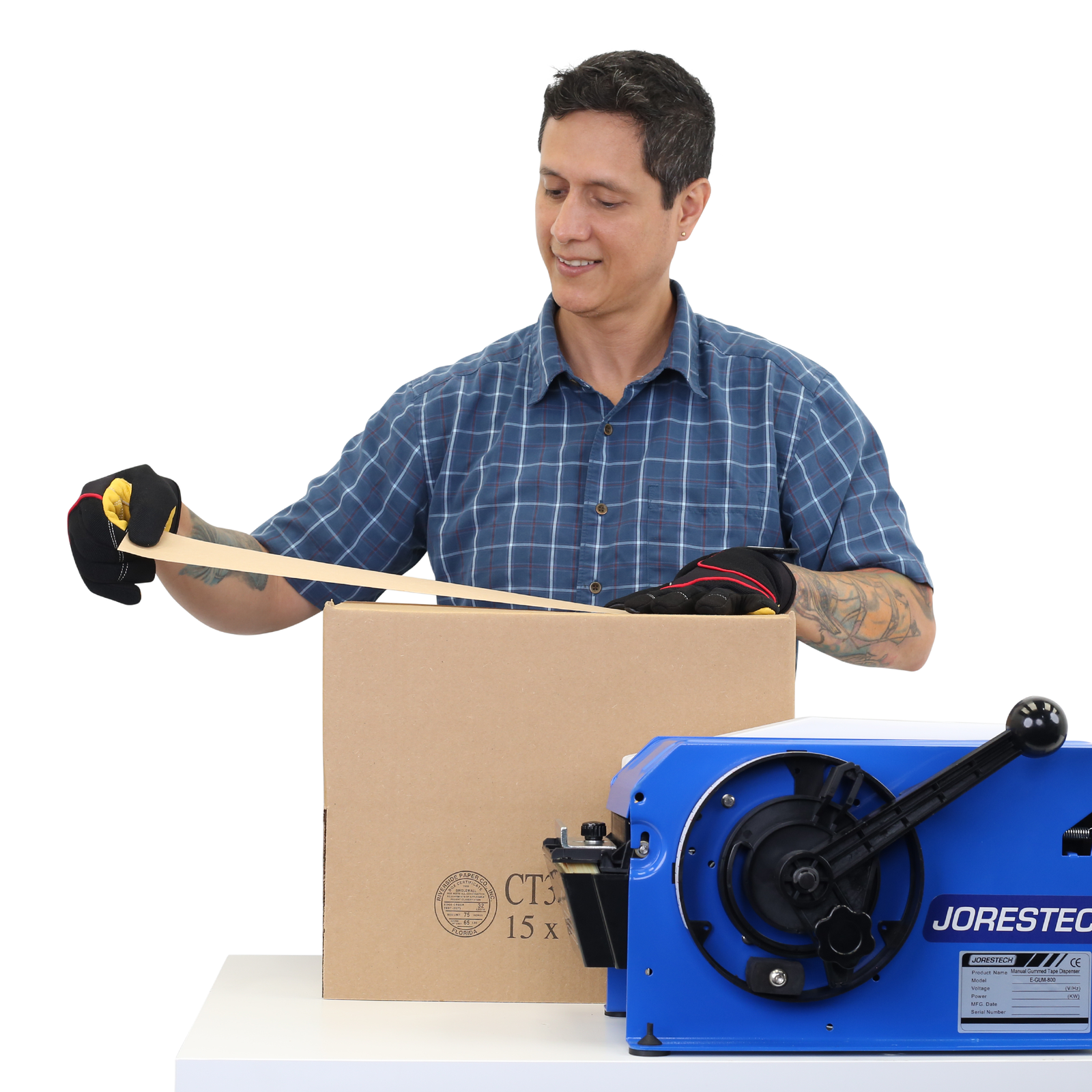 Man placing the wet gummed craft tape on to a brown card box after it was dispensed by the blue JORES TECHNOLOGIES® paper tape dispenser place on the same working table