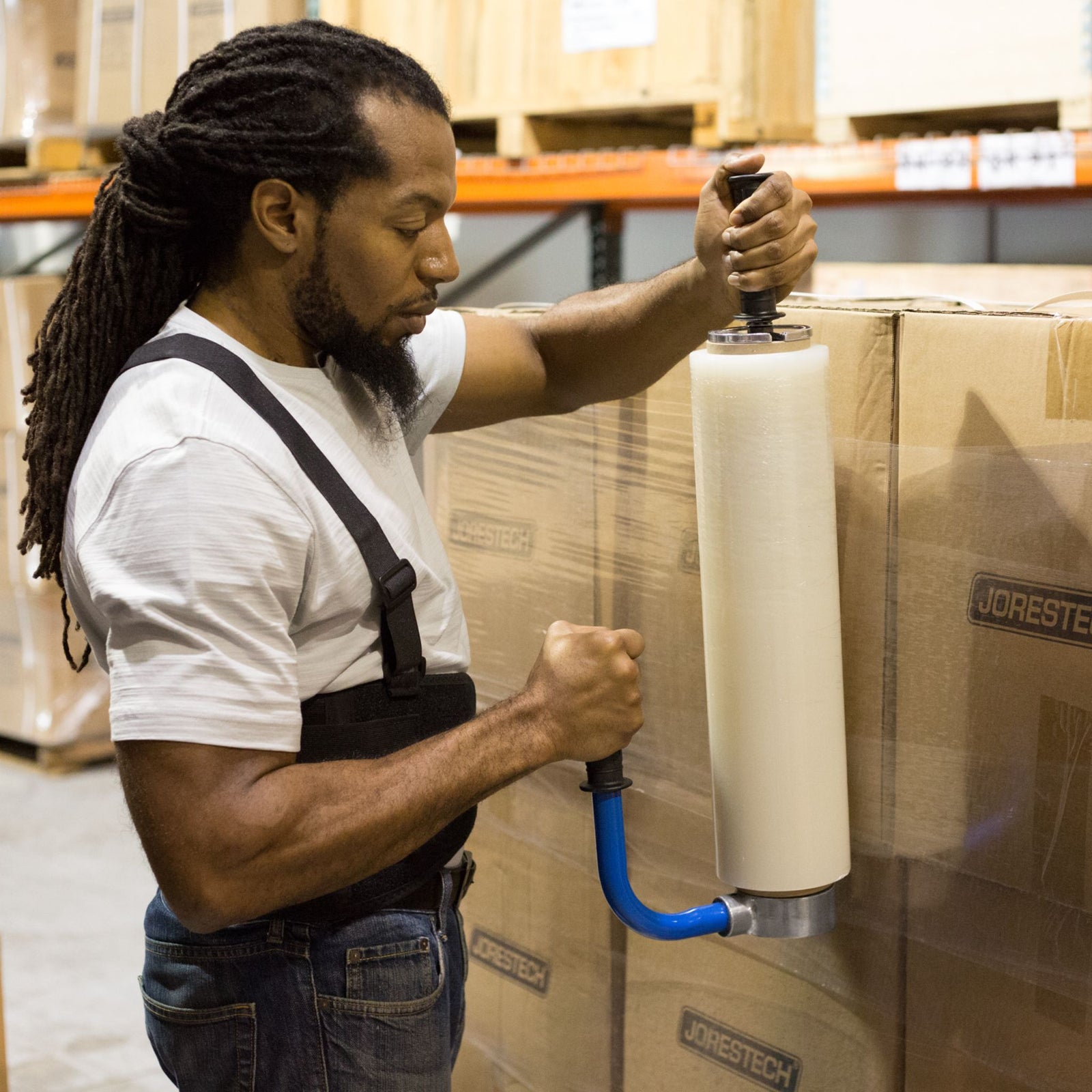 Man in a warehouse wearing a white shirt, jeans and a black back support belt is using the stretch film dispenser to neatly stretch wrap a stack of boxes.