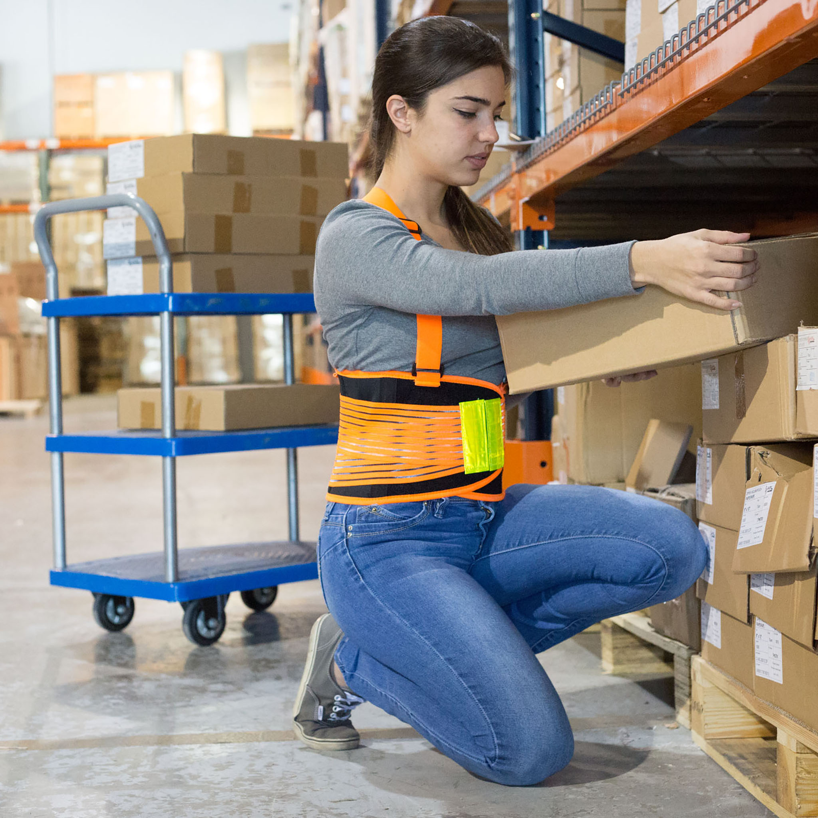 A lady wearing a high visibility adjustable back support belt to avoid back injuries while carrying a heavy box in a warehouse