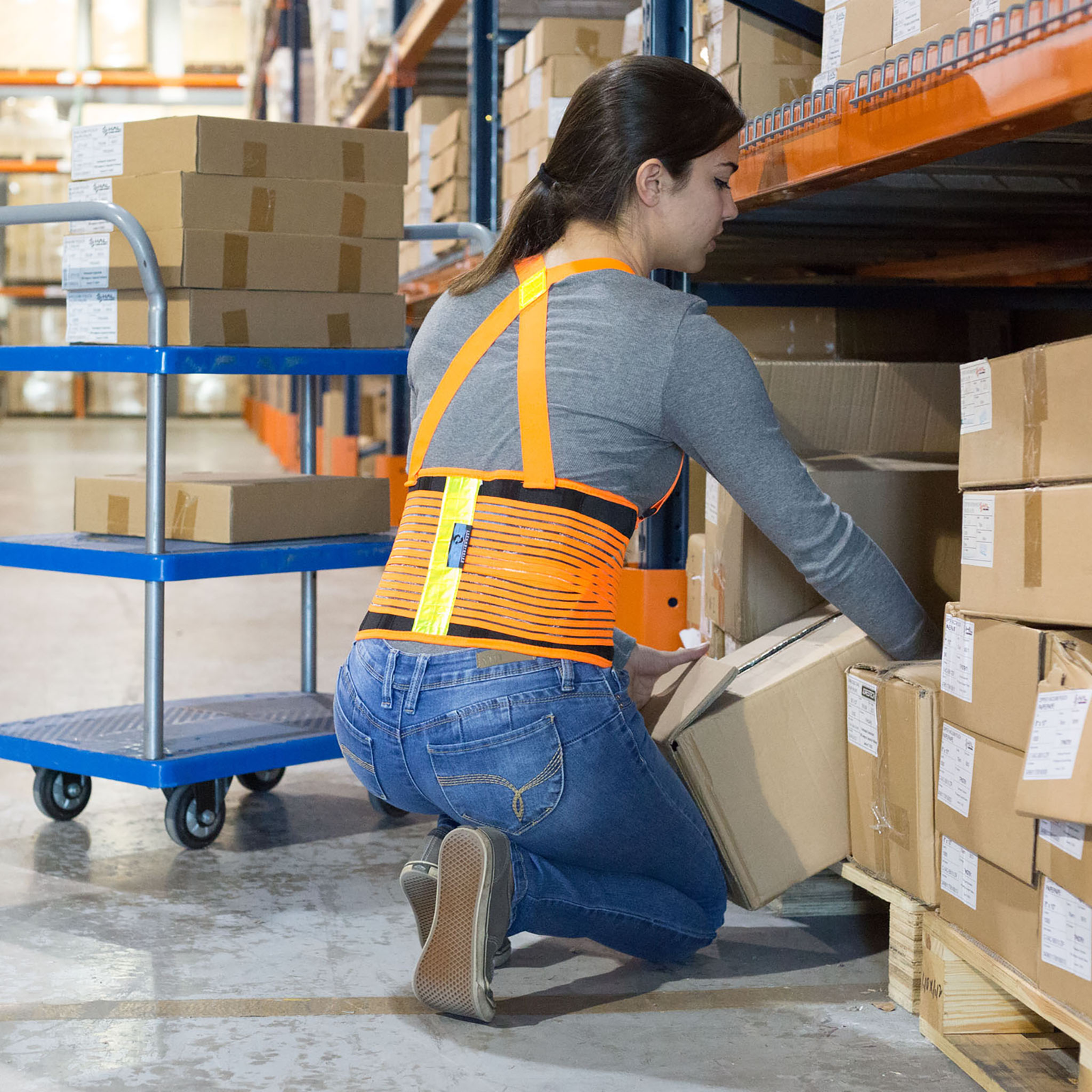 A lady wearing a high visibility adjustable back support belt with reflective stripes while carrying a box in a warehouse. Bright orange color and adjustable straps can be seen in the back.