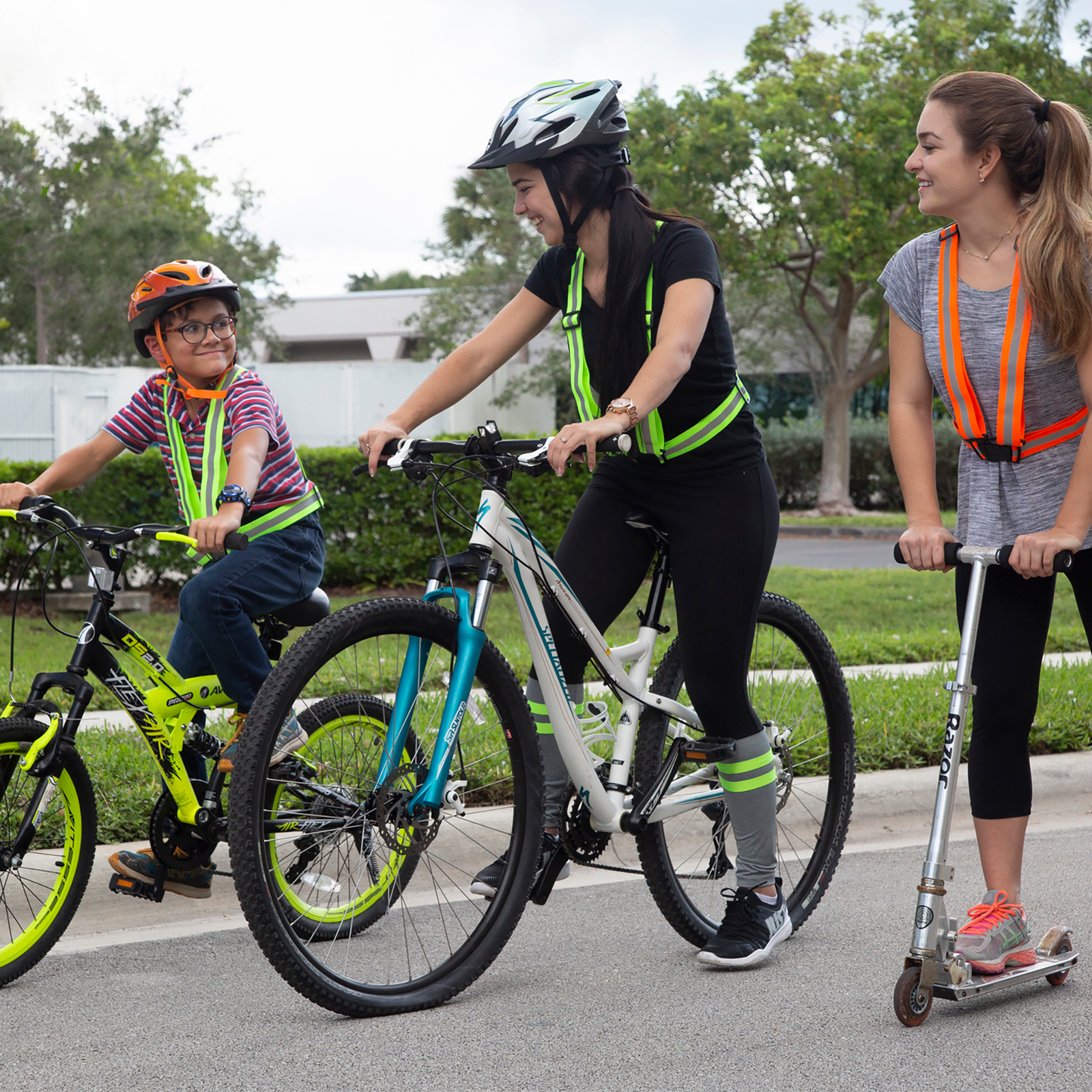 Shows one boy and 2 young ladies riding bike and scooter on the road wearing a the hi visibility safety suspenders for play