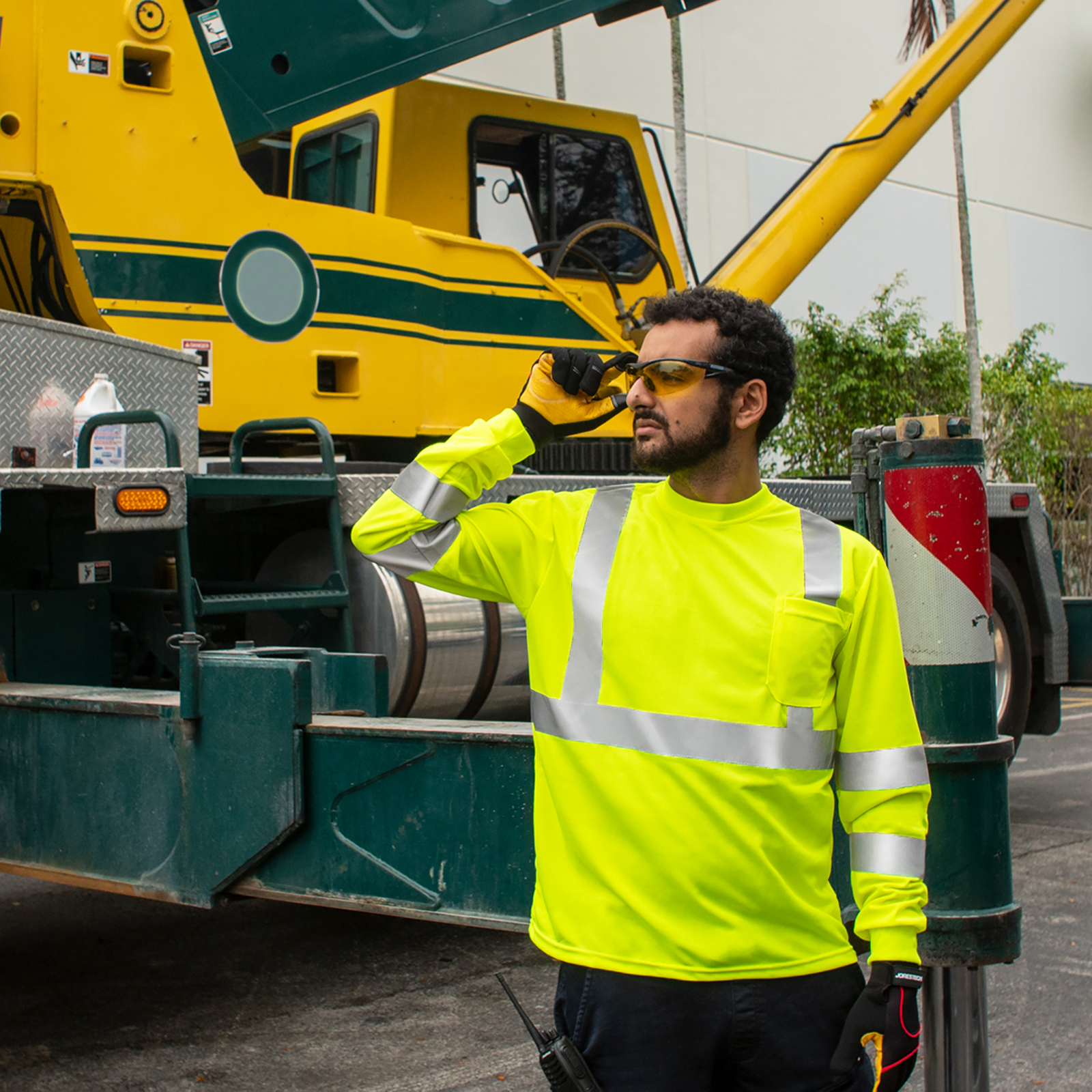 Worker wearing a yellow long sleeve hi vis safety shirt while operating a large green truck in the background