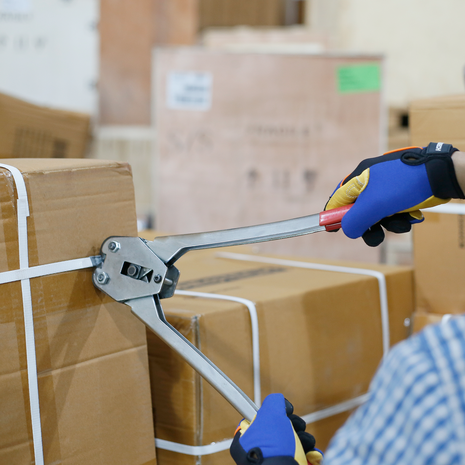 Worker wearing blue and yellow protective gloves. He is using a Manual Poly Strapping Crimper to secure a steel strapping seal on the white poly strap around many carton boxes
