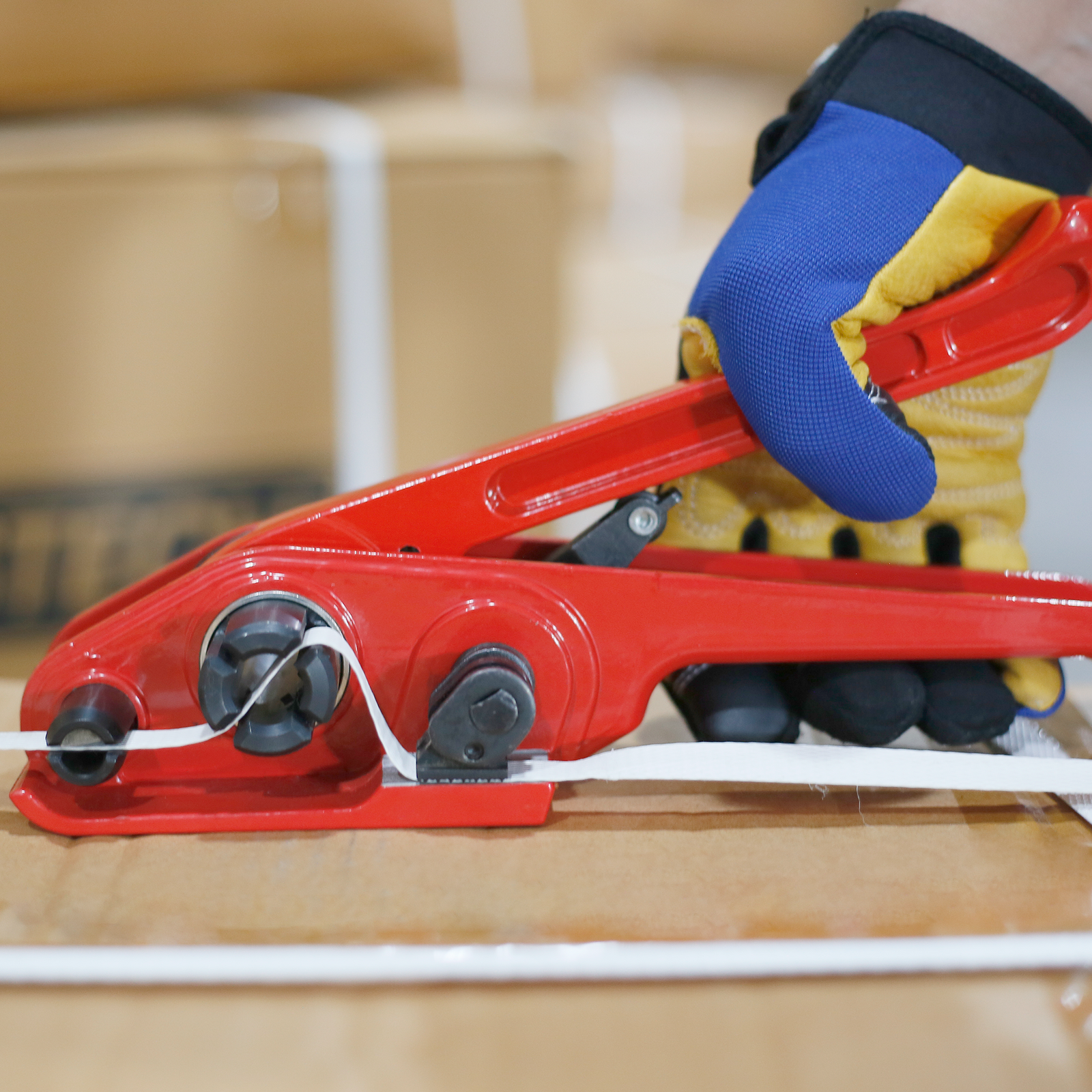 Hand of a person wearing blue mechanical protective gloves holding a Manual Poly Strapping Tensioner threaded with Poly Trap to secure a card box