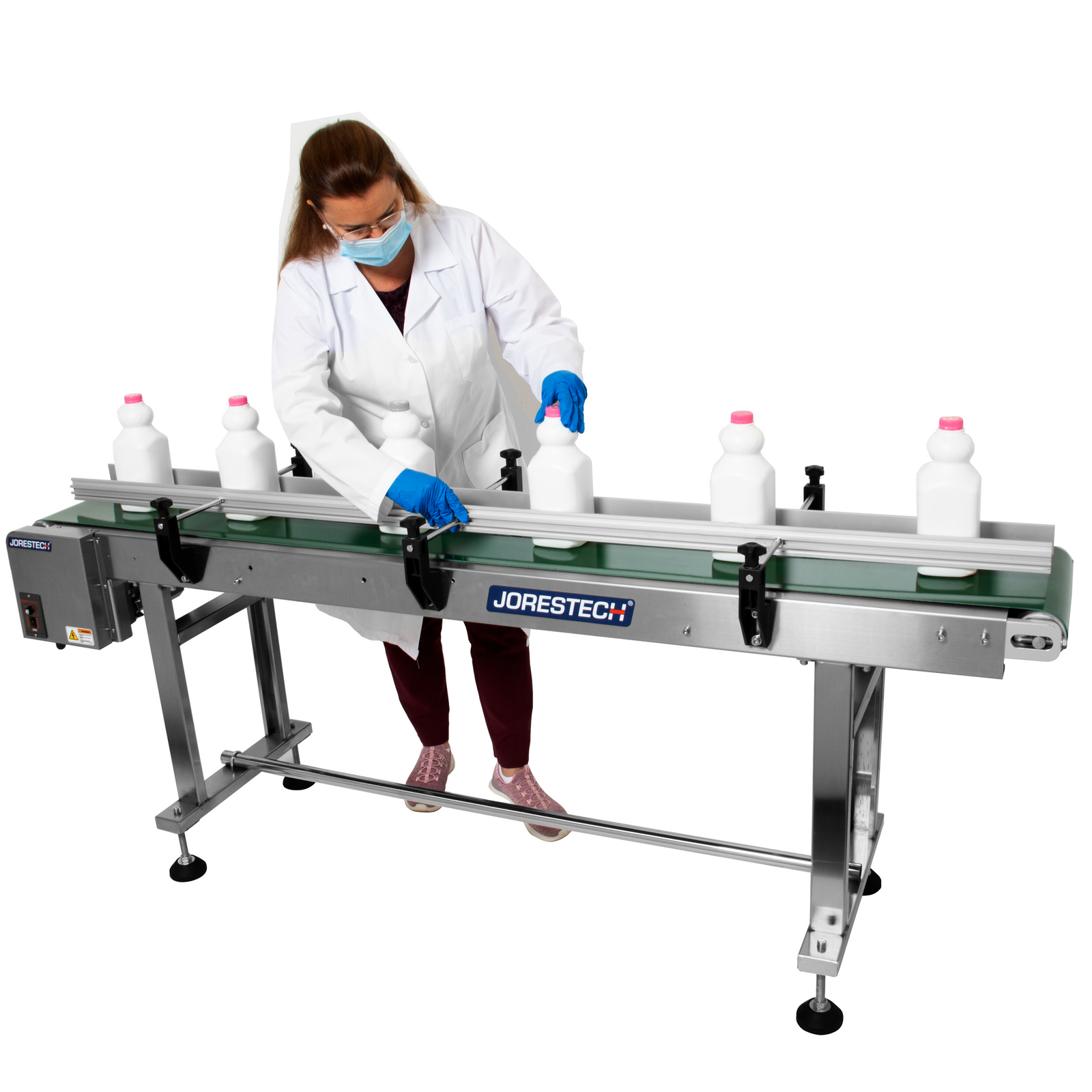 brunette woman wearing white lab coat and blue gloves adjusting white milk bottles on motorized conveyor with steel frame and green belt that features side guard rails