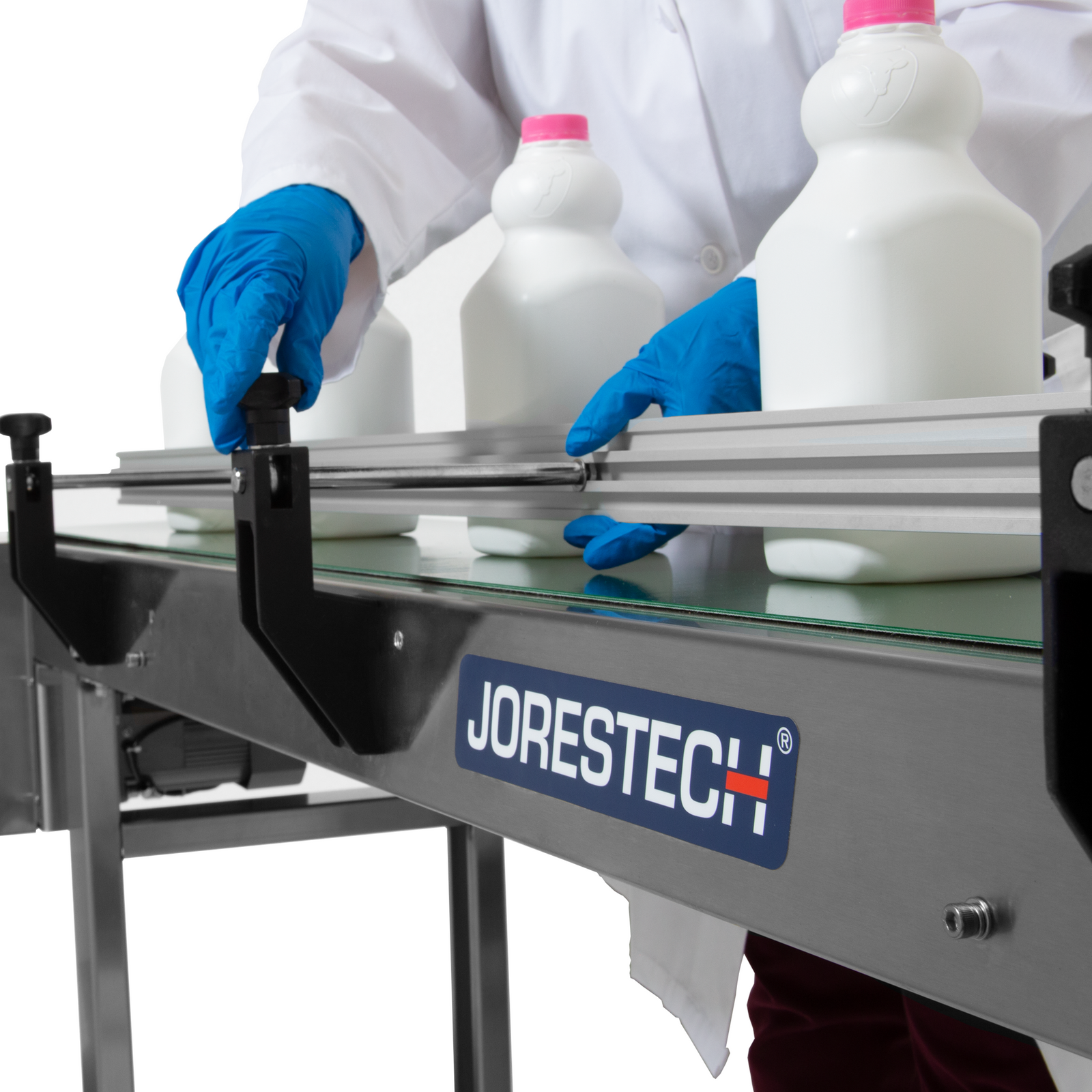 close up of operator wearing white lab coat and blue gloves adjusting white milk bottles on motorized conveyor with steel frame and green belt that features side guard rails.