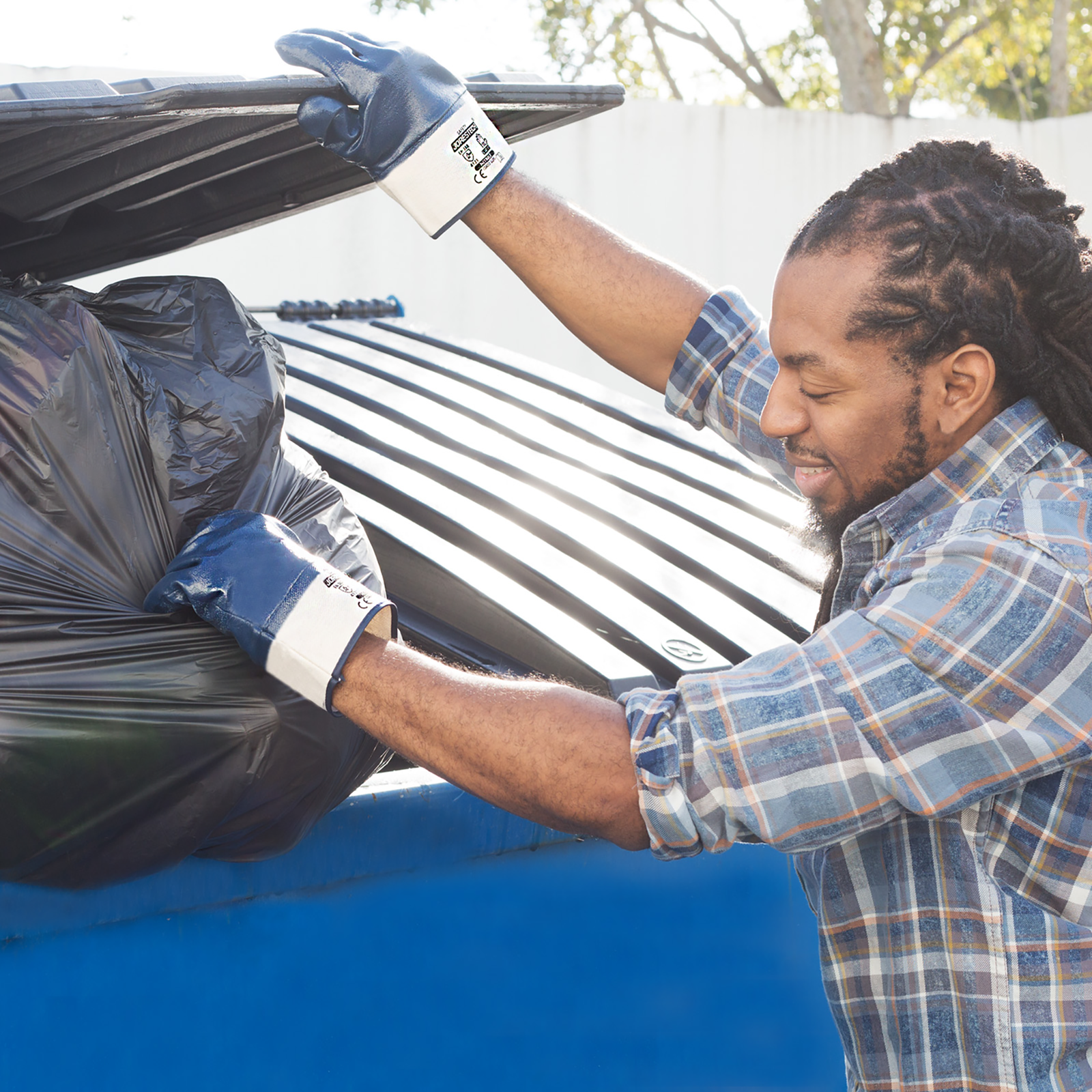Man wearing the JORESTECH nitrile coated safety work gloves for trash removal while putting a black large garbage bag into a dumpster