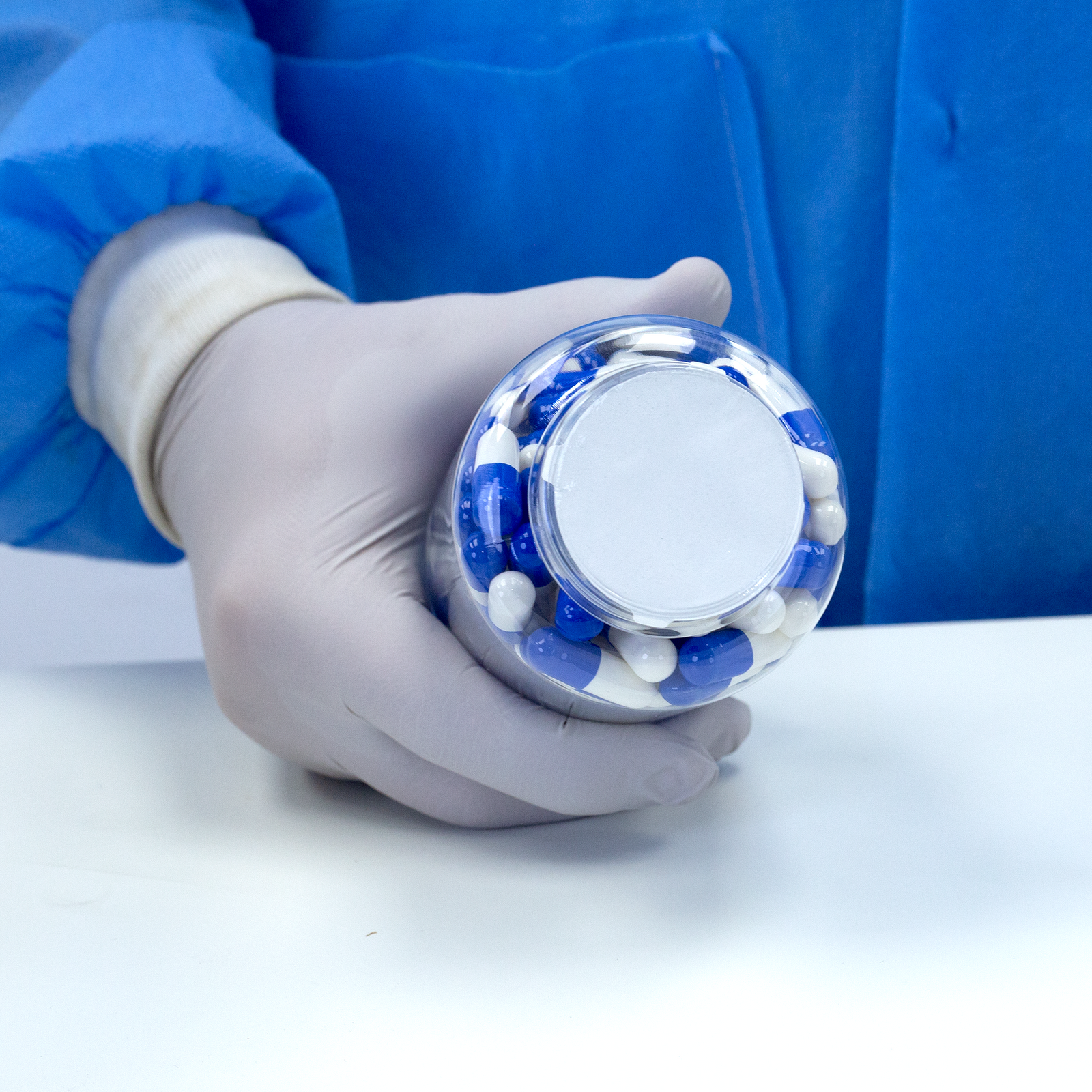Person wearing nitrile gloves and holding a container closed with an induction liner with tabs. Container is filled with blue and white medicine tablets