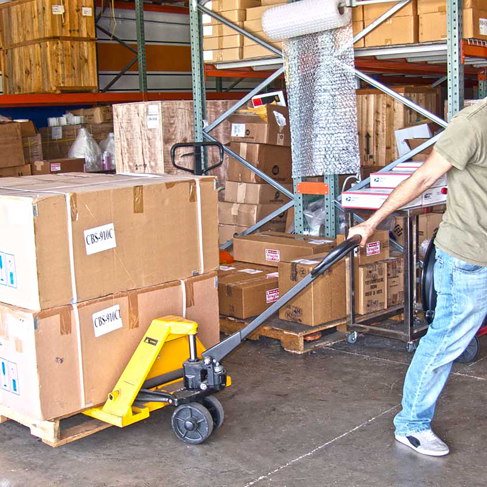 Image shows a person pulling a yellow and black JORESTECH pallet jack truck in a warehouse filled wit boxes.. The pallet jack has several large heavy brown boxes on top. 
