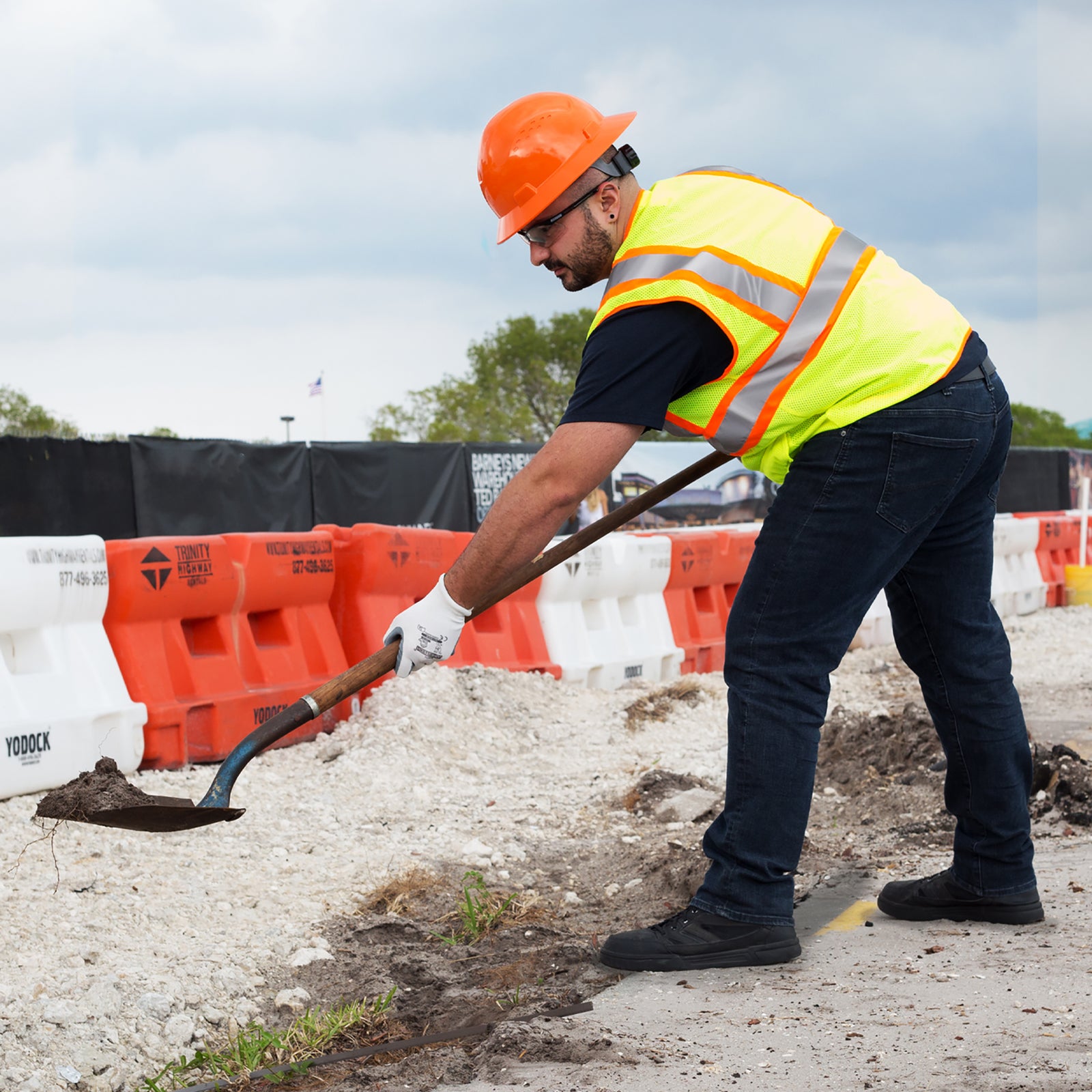 Image of a man wearing a lime orange JORESTECH safety vest, working gloves and an orange hard hat in a construction close to the road