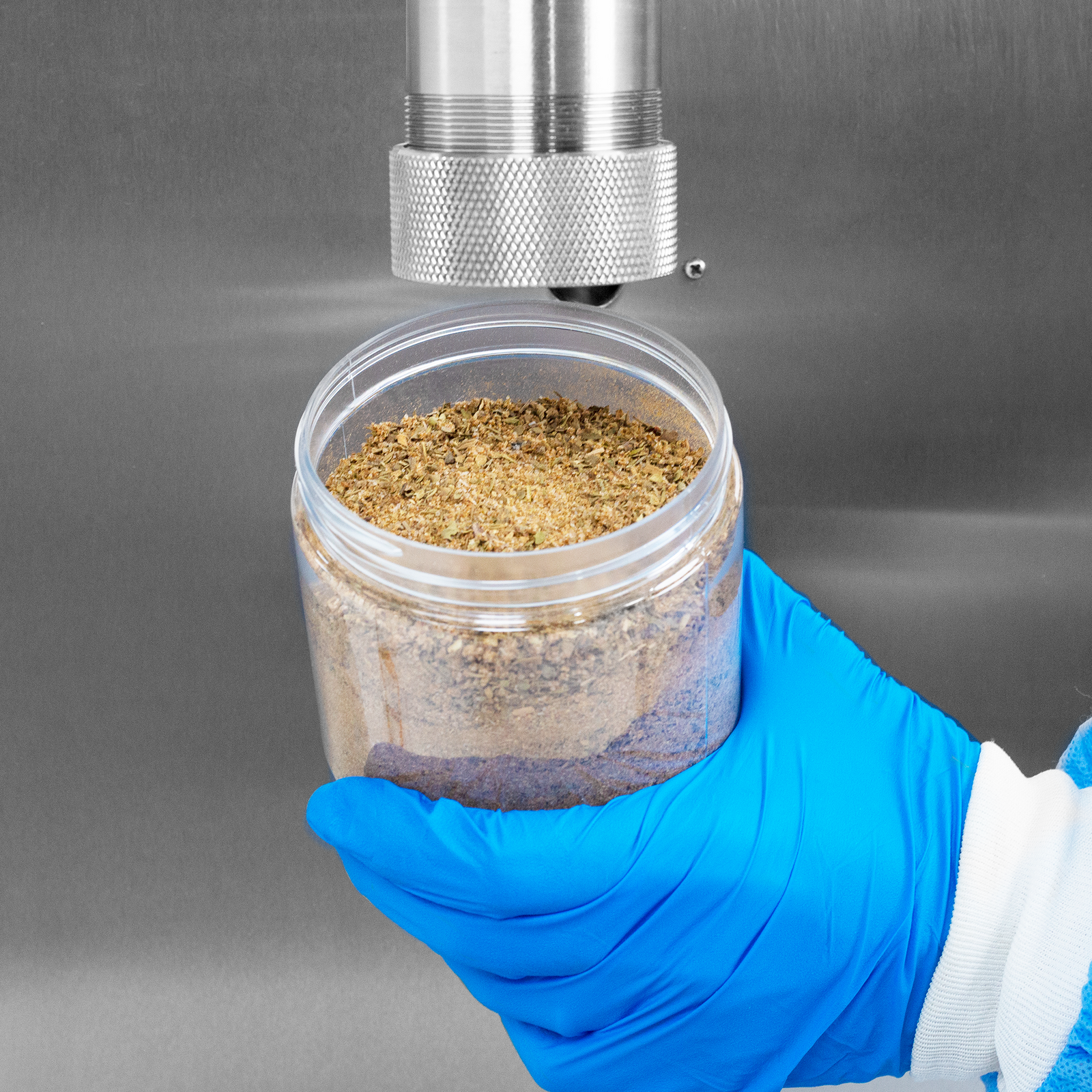 Close-up picture of a person with blue latex gloves and PPE holding a clear container filled with herb seasoning powder. The container is held under the dispensing nozzle of an auger powder filler, which was used to dispense the powder into the container.