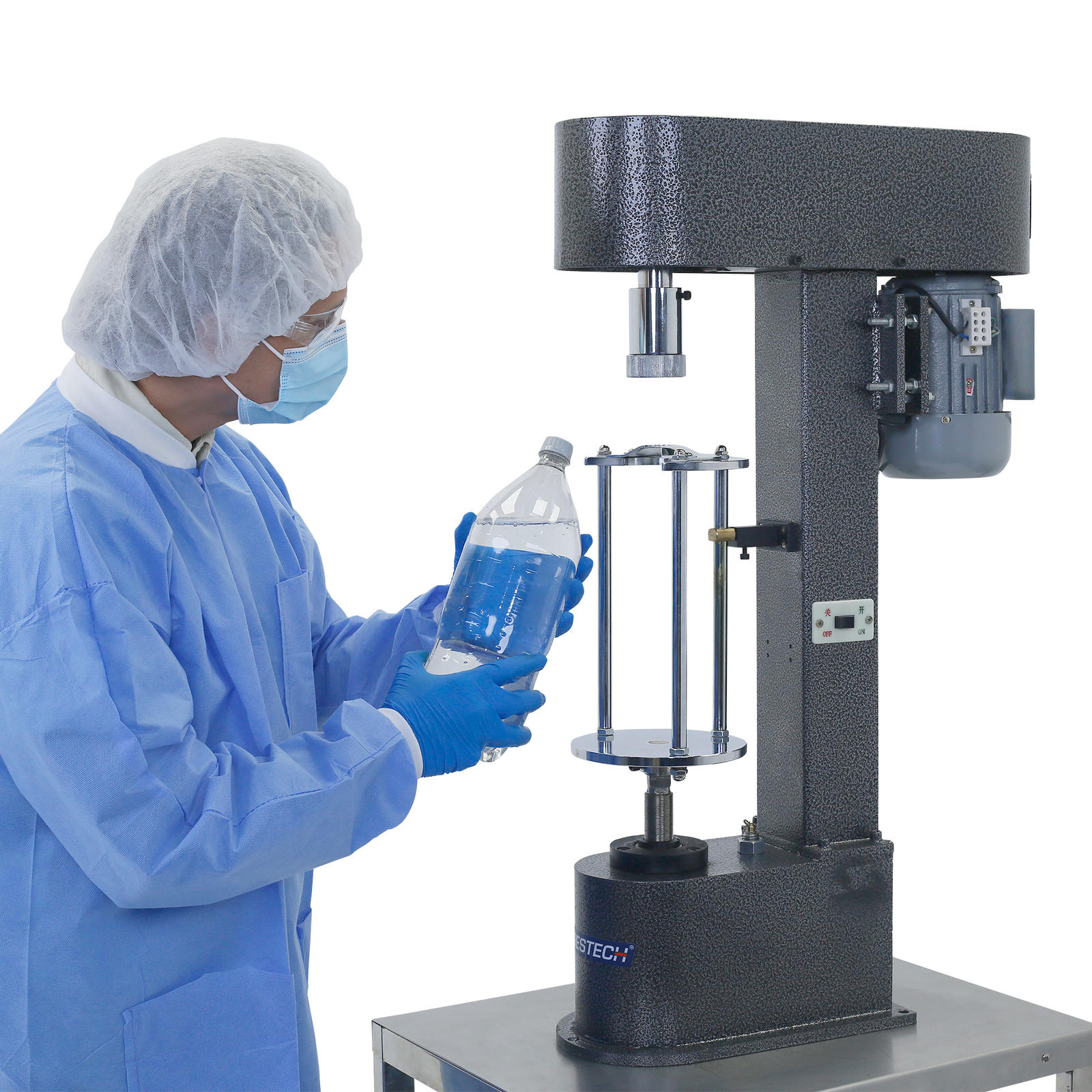 A worker wearing PPE clothing and nitrile gloves is inspecting the plastic cap of a 2 littler plastic clear bottle that was placed by the JORES TECHNOLOGIES® semi-automatic bottle capper for plastic caps. The bottle capper in positioned on top of a working table in front of the worker.
