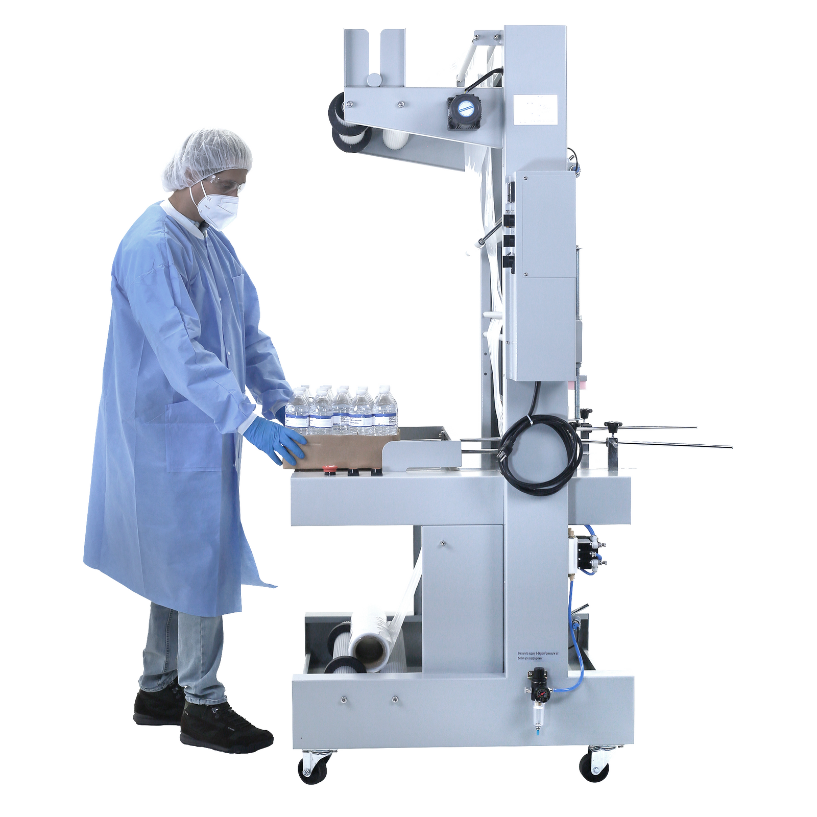 A worker dressed in disposable PPE clothing introducing a card box tray full of water bottles in the JORES TECHNOLOGIES® sleeve shrink sealer