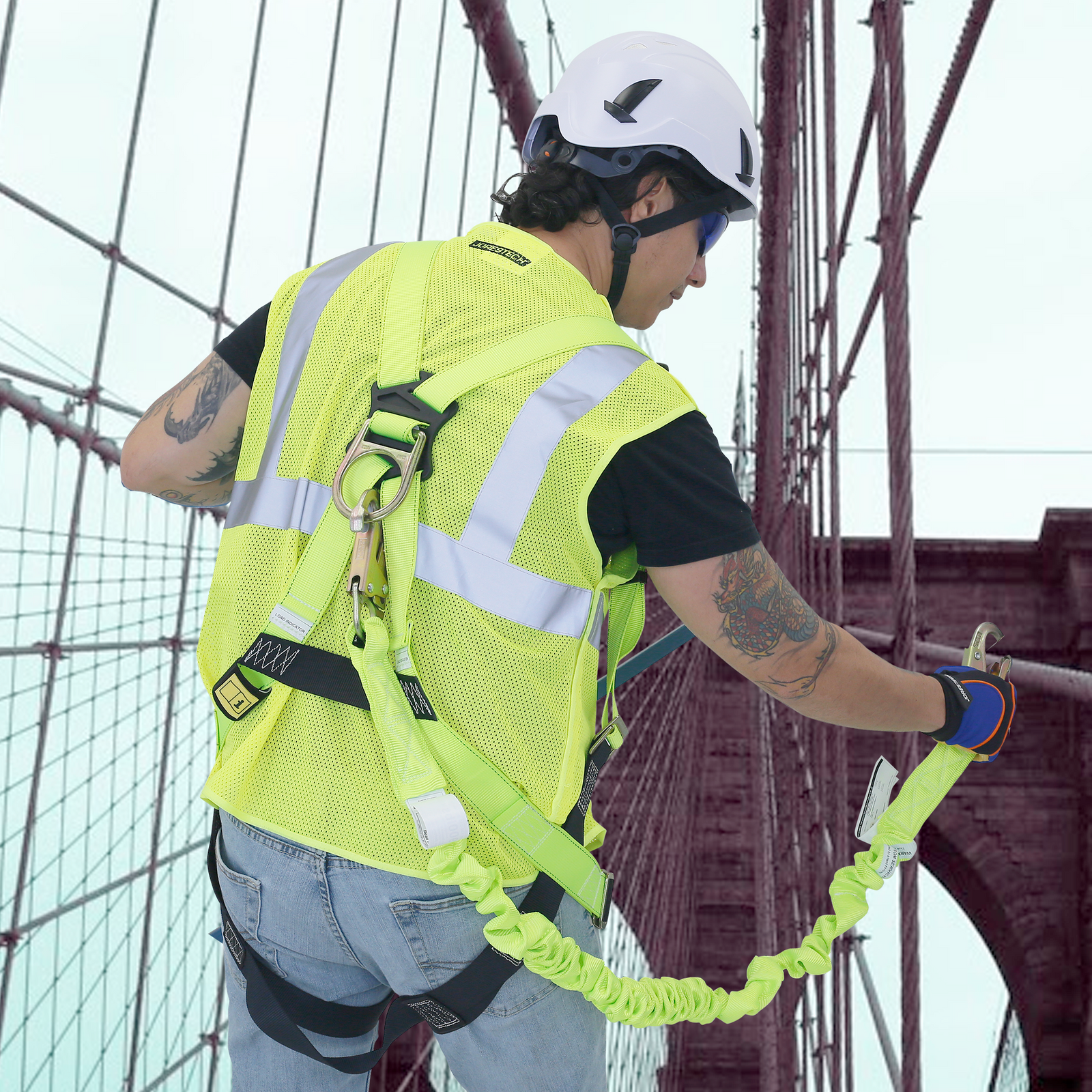 A worker wearing a white hard hat with chin strap and a harness with a single leg internal shock absorbing lanyard hooked to the D ring of the harness. He is securing himself to a stable part of the structure of a bridge while during maintenance or repairs at a very high altitud