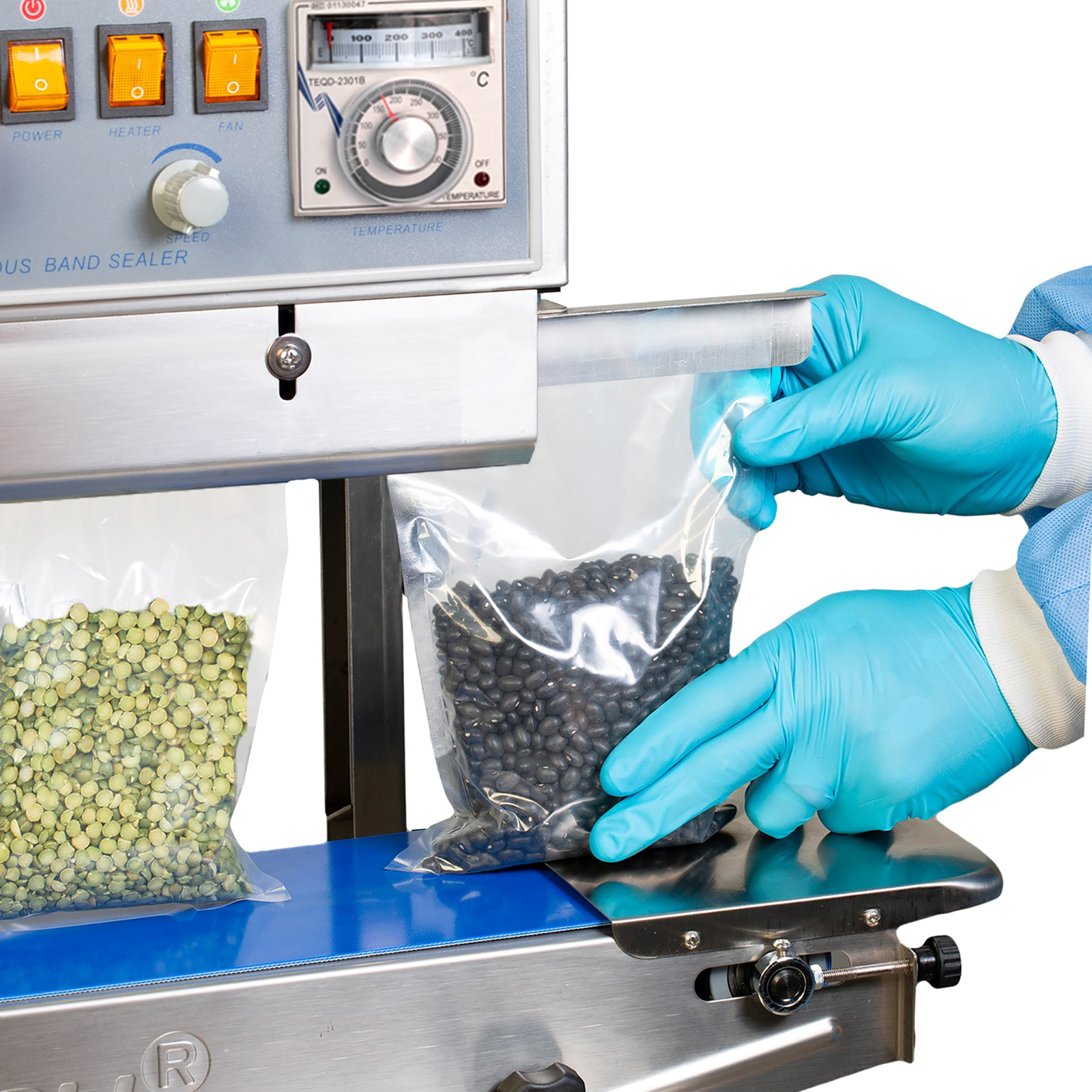 an operator wearing blue gloves while inserting clear bags with black beans and green lentils into a JORESTECH continuous band sealer to close the bags
