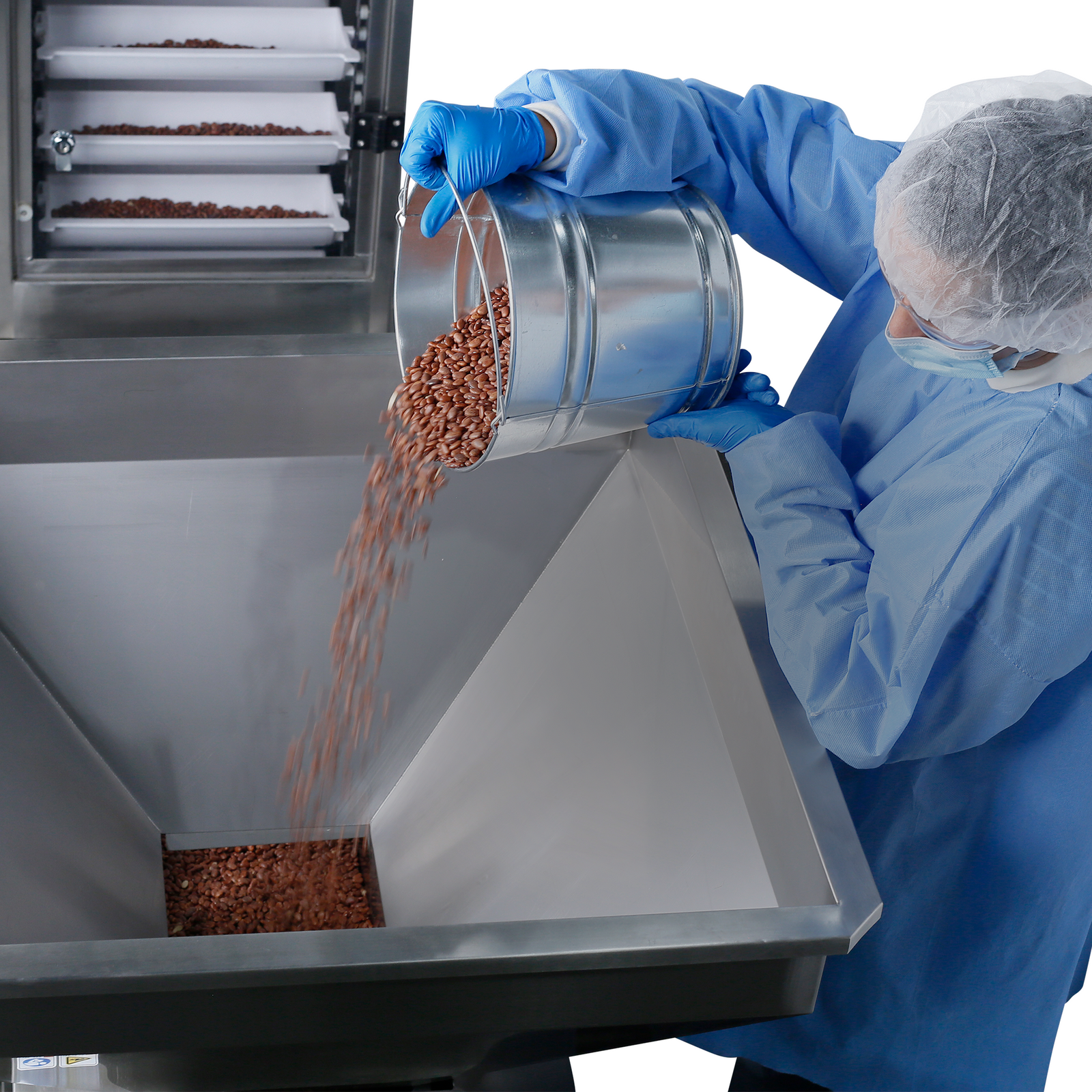 A worker wearing disposable protective equipment dispensing red grains into the hopper of a vibratory feeder positioned next to a JORES TECHNOLOGIES® Motorized Bucket elevator. Beans can be seen transported inside the motorized conveyor