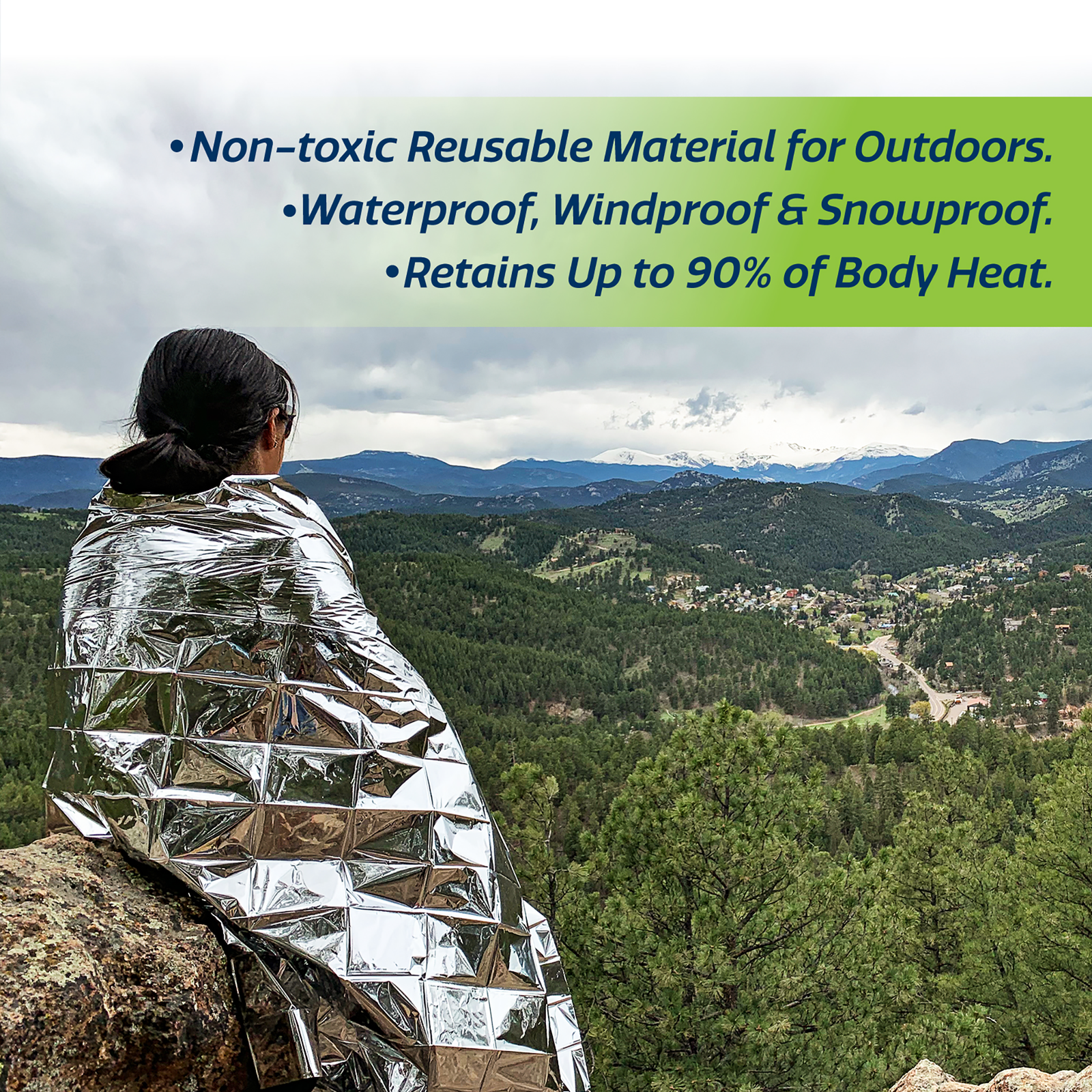 A person covered with a silver color emergency blanket to retain heat while enjoying the view from the top of a mountain. Text reads: Non toxic reusable material for outdoors. waterproof, windproof &snow-proof. retains up to 90% of body heat.