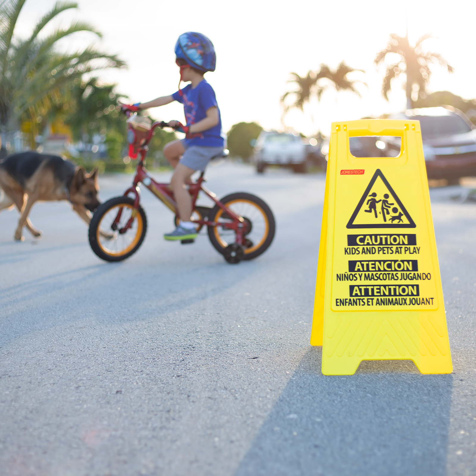 The Jorestech two sided folding kids at play caution stand. There is a little kid riding his bicycle with his dog next to him on the background