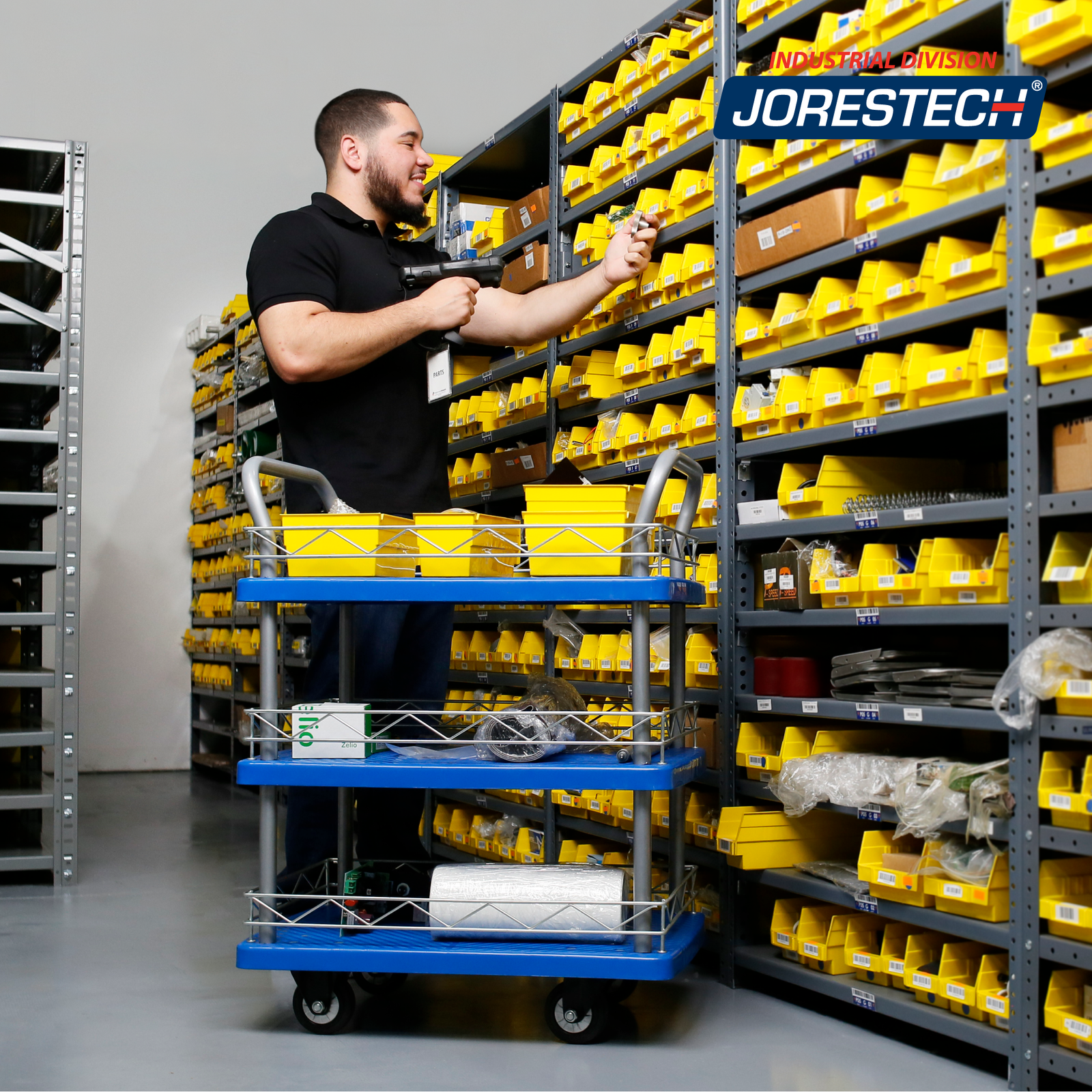 A man in a warehouse scanning some items. He is using the JORES TECHNOLOGIES®  3 shelf utility push cart to roll his items around the workplace